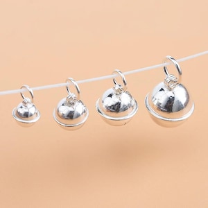 Puede incluir: Cuatro cascabeles esféricos de color plateado de diferentes tamaños, cada uno con una pequeña argolla circular en la parte superior. Los cascabeles están dispuestos en fila, suspendidos de un hilo blanco fino, mostrando un acabado metálico liso y brillante.