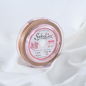 May include: A spool of pink gold nylon coated wire, labeled "SakuLux." The spool is encased in a clear plastic container with pink accents. The label indicates a 0.40 mm coated diameter and 100m length. The wire is for jewelry making.