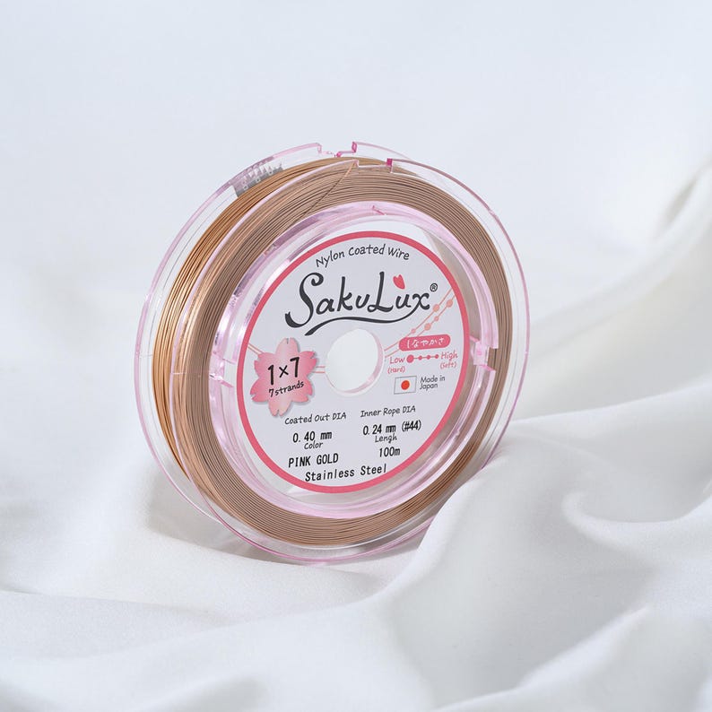 May include: A spool of pink gold nylon coated wire, labeled "SakuLux." The spool is encased in a clear plastic container with pink accents. The label indicates a 0.40 mm coated diameter and 100m length. The wire is for jewelry making.