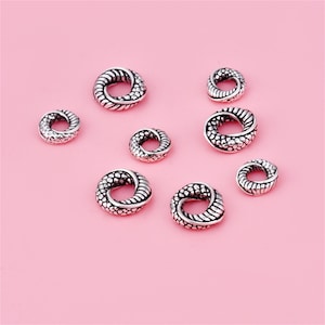 May include: A selection of silver-toned, circular beads with a textured, braided design. The beads are displayed on a pink background. The beads vary in size, with a detailed pattern on the surface.