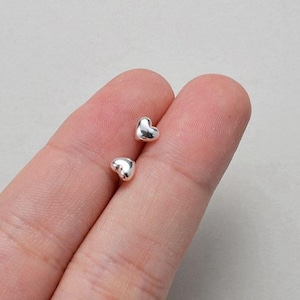 Könnte beinhalten: Zwei kleine, silberne herzförmige Ohrstecker. Die Ohrringe sind glatt und glänzend und reflektieren das Licht. Die Ohrringe werden auf einem hellen Hintergrund präsentiert.
