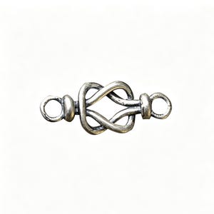 May include: A silver-coloured jewellery clasp in the shape of a Celtic knot. The clasp features a central knot design with loops at each end for attachment. The metal has a slightly aged appearance.