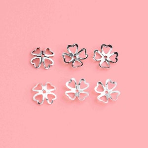 May include: Six silver flower-shaped bead caps on a pink background. The caps have a delicate, openwork design.