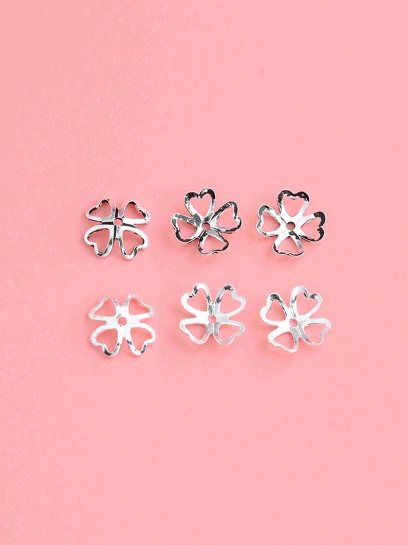 May include: Six silver flower-shaped bead caps on a pink background. The caps have a delicate, openwork design.