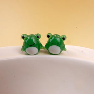 May include: Two small, green frog figurines with black eyes and white bellies. The frogs are smiling and appear to be made of a translucent material. They are sitting on a white surface against a yellow background.