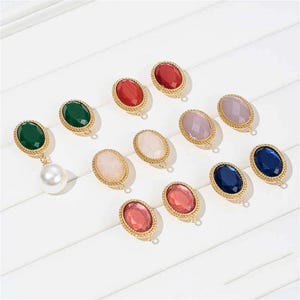Puede incluir: Colección de pendientes ovalados con piedras preciosas de varios colores, incluyendo verde, rojo, rosa y azul, con engastes de metal dorado. Un pendiente presenta una gran perla blanca colgante.