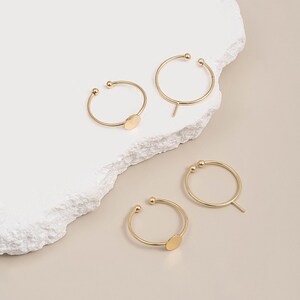 May include: Four gold-colored adjustable rings. Two rings have a small, flat, oval-shaped accent. The other two rings have a small ball on each end and a small, straight, vertical bar. The rings are on a white, textured surface.