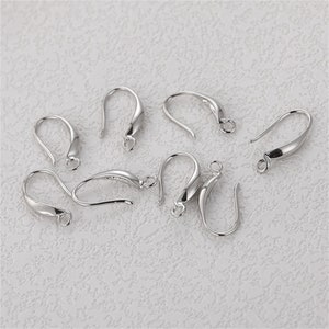 May include: A collection of silver-colored earring hooks. The hooks have a curved design with a small loop at the bottom and a rounded top. The hooks are arranged on a textured, off-white surface.