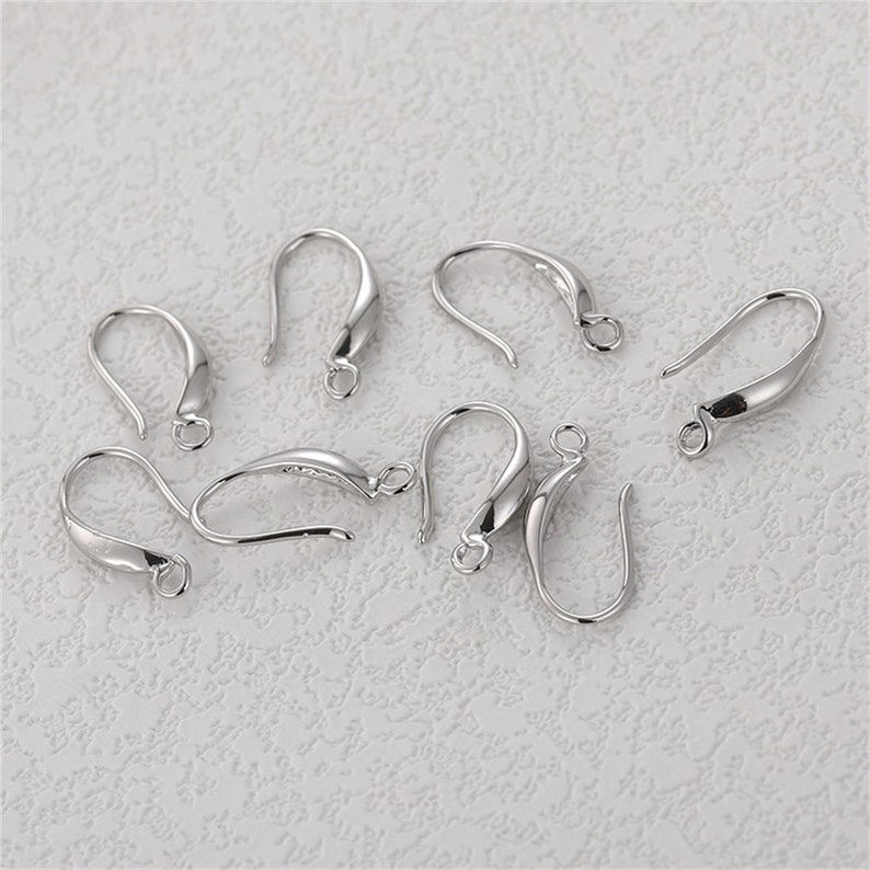 May include: A collection of silver-colored earring hooks. The hooks have a curved design with a small loop at the bottom and a rounded top. The hooks are arranged on a textured, off-white surface.