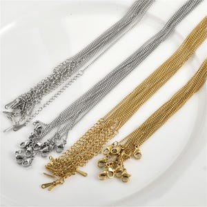 Könnte beinhalten: Nahaufnahme von mehreren silber- und goldfarbenen Kettenhalsketten mit Karabinerverschlüssen. Die Ketten sind in ordentlichen Reihen angeordnet, wodurch ihre filigranen Glieder und das glänzende Finish hervorgehoben werden. Die Halsketten werden auf einer weißen Oberfläche präsentiert.