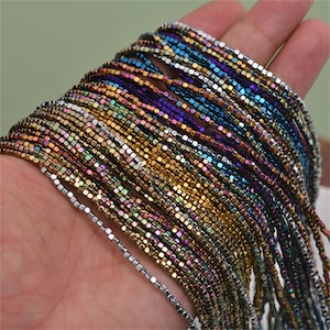 May include: A strand of iridescent square beads in various colours, including silver, gold, blue, purple, and green. The beads are small and uniform in size, creating a shimmering effect.