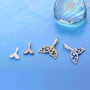 May include: Four different mermaid tail pendants in silver, rose gold, and gold tones, each adorned with sparkling clear crystals. The pendants are arranged on a light blue surface, with a decorative glass stone in the background.