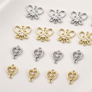 May include: A collection of metal jewelry charms featuring silver and gold-toned butterfly and heart shapes. Each charm is accented with a single clear, round-cut cubic zirconia stone and includes small loops at the top and bottom for attachment.