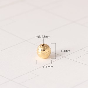 May include: A gold-coloured round bead with a textured surface and a hole in the centre. The bead measures 5.3 mm in diameter and 5.5 mm in height. The hole is 1.3 mm in diameter.