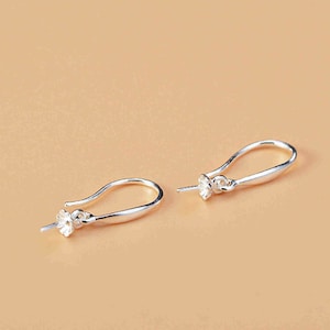 Può includere: Due ganci per orecchini color argento con un design floreale. I ganci hanno una forma curva e allungata, e i dettagli floreali sembrano essere fatti di piccole pietre scintillanti. Gli orecchini sono esposti su uno sfondo marrone chiaro.