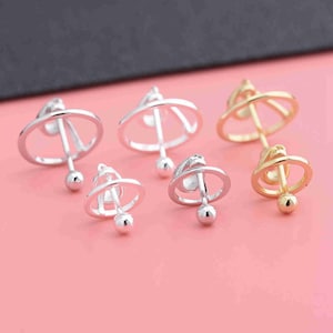Puede incluir: Cinco pendientes giratorios de plata y oro con un diseño circular. Los pendientes están sobre una superficie rosa.