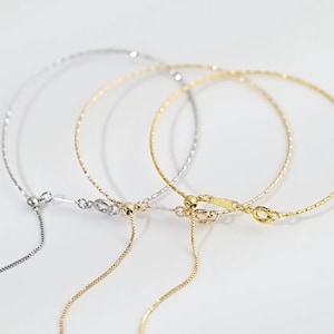 Könnte beinhalten: Drei zarte Halsketten in Silber- und Goldtönen. Jede Halskette hat eine dünne, strukturierte Kette und einen verstellbaren Verschluss. Die Halsketten werden vor einem weißen Hintergrund präsentiert, was ihr minimalistisches Design und ihre feinen Details hervorhebt.