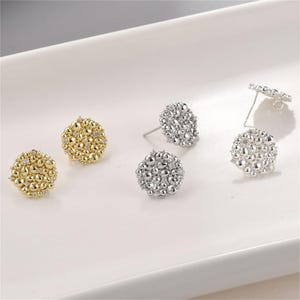 May include: A collection of stud earrings in gold and silver tones. The earrings have a textured, clustered design, with some featuring small, clear accents. The earrings are displayed on a white surface, showcasing their intricate details.