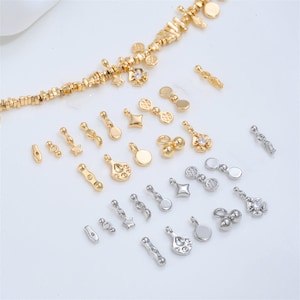 May include: A collection of small metal jewelry charms in gold and silver tones, featuring various shapes including stars, circles, teardrops, and geometric designs, some accented with clear crystals, arranged alongside a beaded chain.