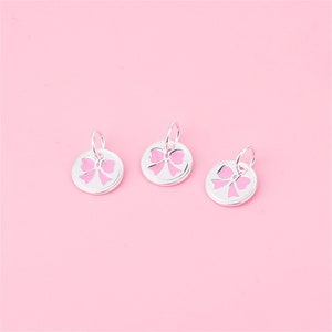 May include: Three silver charms with a pink bow cut-out design. The charms are round and have a loop at the top for attaching to a bracelet or necklace.