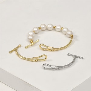 Puede incluir: Dos pulseras de oro y plata con cierre de palanca. Cada pulsera tiene una forma curva irregular y está adornada con una cadena de perlas blancas.