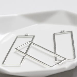Puede incluir: Marcos rectangulares plateados con un lazo en la parte superior para la fabricación de joyas. El diseño de marco abierto se muestra sobre una superficie blanca, mostrando las formas geométricas simples. Ideal para crear pendientes o collares.