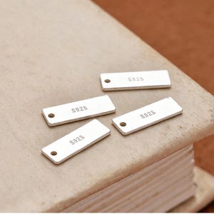 May include: Four rectangular silver tags with rounded corners and a small hole on one end. Each tag has the text "S925" printed on it. The tags are lying on a textured, light brown surface.