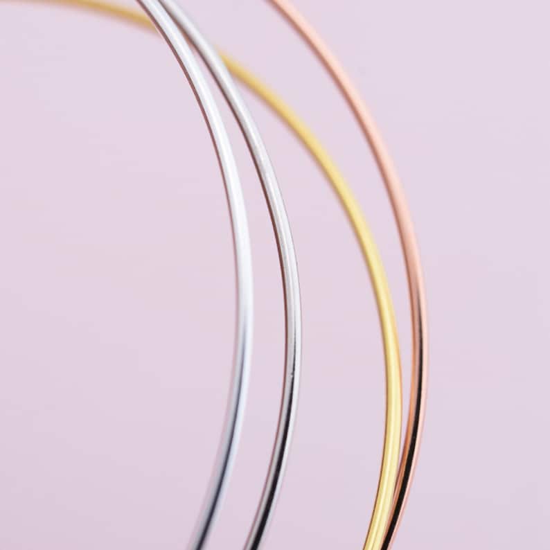 May include: Three thin metal hoops in silver, gold, and rose gold, arranged in a circle on a pink background.