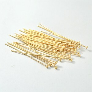 Puede incluir: Un mont&oacute;n de alfileres de cabeza dorados con extremos redondeados. Los alfileres est&aacute;n dispuestos en un patr&oacute;n aleatorio sobre una superficie blanca.