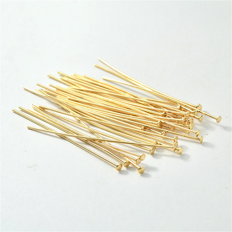 Puede incluir: Un mont&oacute;n de alfileres de cabeza dorados con extremos redondeados. Los alfileres est&aacute;n dispuestos en un patr&oacute;n aleatorio sobre una superficie blanca.
