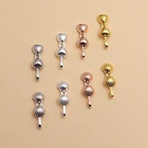 May include: A selection of metal jewellery clasps in silver, rose gold, and gold hues. The clasps showcase heart and seashell designs, each with a small hook. The clasps are presented against a neutral beige backdrop.