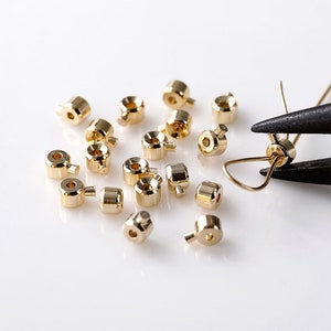 May include: A group of gold-colored metal beads with a small hole in the center of each bead. The beads are scattered on a white surface.