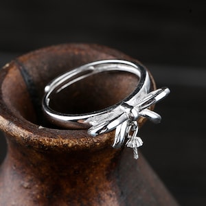 Può includere: Un anello color argento con un design a libellula. L'anello presenta una libellula dettagliata con ali spiegate e un piccolo ciondolo pendente. L'anello è ambientato su uno sfondo scuro e sfocato.
