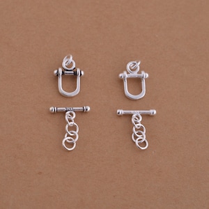 Könnte beinhalten: Zwei silberfarbene Toggle-Verschlüsse mit einem Kettenverbinder. Jeder Verschluss hat einen D-förmigen Ring und eine T-förmige Stange mit einer kleinen Kugel an jedem Ende.