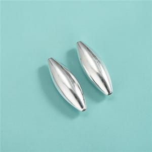 Pode incluir: Duas contas alongadas polidas, de cor prateada, com formato oval simétrico. As contas estão sobre um fundo azul-petróleo claro, criando um contraste que realça suas superfícies refletoras. As contas são provavelmente para fazer joias.