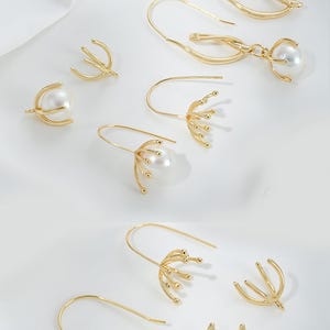 May include: A set of gold-tone earring findings with pearl accents. The findings feature different shapes and designs, including a simple hoop, a curved design, and a delicate floral design.