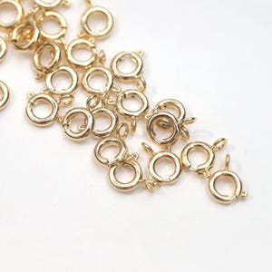 May include: Close-up of several gold-colored spring ring clasps, a jewelry-making component. The clasps are round with a small lever to open and close. They are scattered on a white surface, ideal for jewelry making.