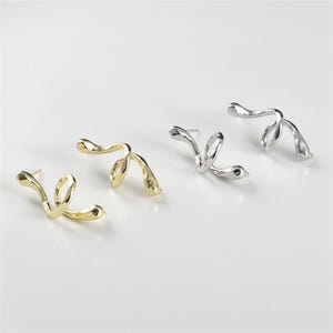 Könnte beinhalten: Zwei Paar Ohrringe, eines golden und eines silbern, in Form einer Schlange. Die Ohrringe sind aus glattem, strukturiertem Metall gefertigt und haben ein leicht abstraktes Design.