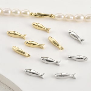May include: Gold and silver fish-shaped jewellery components. The gold fish have a shiny finish, while the silver ones are polished. A pearl necklace with a gold fish-shaped clasp is also visible. These components are likely for jewellery making.