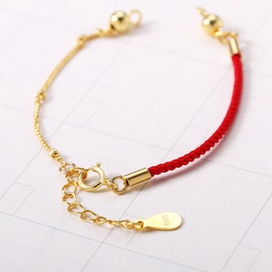 May include: A gold-toned bracelet with a red cord section. The bracelet features a delicate chain and a clasp with an extender. A small tag with "S925" is attached. The bracelet is displayed on a white surface.