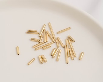 May include: A close-up of a white plate with a scattering of gold metal tube beads. The beads are various lengths and are arranged in a random pattern.