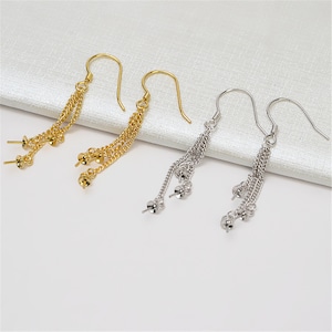 May include: Gold and silver-colored dangle earrings with multiple chains and small bead accents. The earrings have a hook design for easy wear. The jewelry is displayed on a white, textured surface.