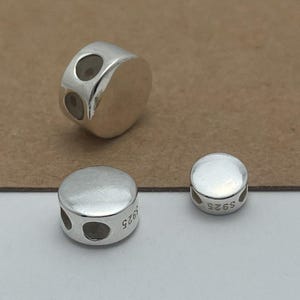 Pode incluir: Três contas de prata polida com um topo plano e circular e um perfil lateral arredondado. Cada conta apresenta dois pequenos orifícios circulares na lateral para enfiar. A lateral das contas está claramente marcada com o texto S925 em preto.