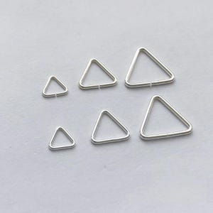 May include: Six silver triangle earrings of different sizes are displayed on a light grey background. The earrings are simple geometric shapes, with a small gap at one point of the triangle.