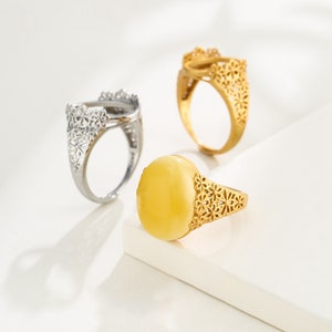 Puede incluir: Tres anillos con intrincados diseños florales. Dos anillos son de oro y uno es de plata. El anillo de oro de la derecha tiene una piedra ovalada amarilla.