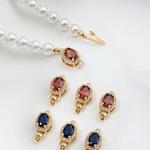 Op de afbeelding: Een set gouden sluitingen voor kettingen of armbanden, met ovale rode en blauwe edelstenen omringd door kleinere heldere stenen.