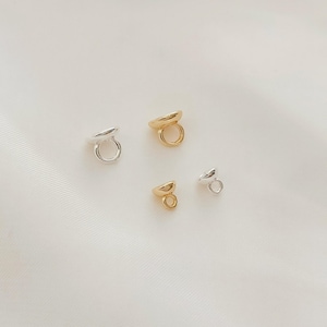 Puede incluir: Cuatro pequeños hallazgos de joyería en tonos dorados y plateados están dispuestos sobre una superficie blanca. Cada hallazgo tiene una parte superior redondeada y un pequeño lazo. Probablemente se utilizan para la fabricación de joyas.