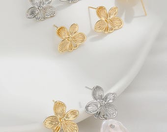 May include: A collection of flower-shaped earrings in gold and silver tones. Some earrings are simple studs, while others feature a large, irregular white pearl drop. The earrings are displayed on a white surface.