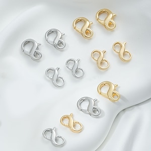 May include: Assortment of silver and gold infinity shaped lobster clasps for jewelry making. The clasps are arranged in two rows on a white surface.
