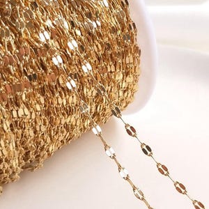 Könnte beinhalten: Eine Spule mit goldfarbener Kette mit einem filigranen, flachen, oval geformten Kettenglied-Design. Die Kette ist eng gewickelt, wobei ein Teil herausragt, um die einzelnen Glieder zu zeigen. Die Kette hat eine glänzende, reflektierende Oberfläche.